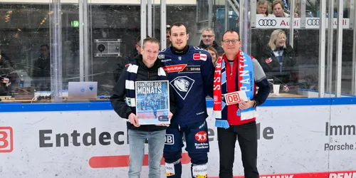 Ein Eishockeyspieler steht zwischen zwei weiteren Personen auf einer Eisfläche und bekommt einen Preis überreicht
