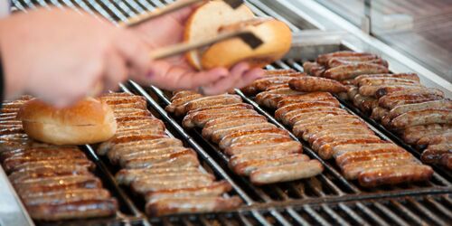 Bratwürste auf dem Grill mit Händen im Vordergrund, die eine Wurst in ein Brötchen legen